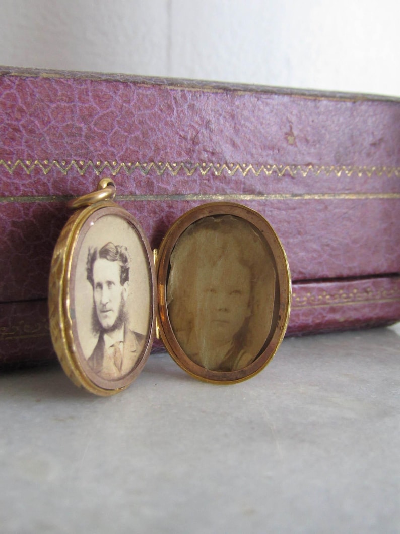 Op de afbeelding: Open gouden medaillon met twee ovale compartimenten, elk met een sepia-getint portret. E&eacute;n toont een man in pak en baard. Het medaillon ligt in de buurt van een paarse, getextureerde doos.