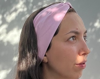 Può includere: Una donna con lunghi capelli castani indossa una fascia per capelli annodata di colore rosa chiaro. Sta guardando a destra e ha un'espressione neutra sul viso.