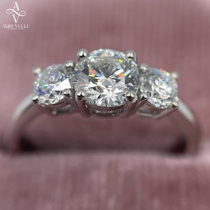 May include: A three-stone ring featuring a large round brilliant-cut centre diamond flanked by two smaller round diamonds, all set in a polished silver-toned metal band. The Auri Velle logo is visible in the upper left corner of the image.