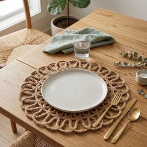 Könnte beinhalten: Ein rundes, beiges Platzset aus geflochtener Jute mit einem dekorativen Rand aus Schlaufen. In der Mitte des Platzsets steht ein schlichter, matter weißer Keramikteller. Daneben liegen eine goldfarbene Gabel, ein Messer und ein Löffel aus Metall.