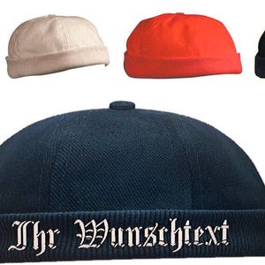 Könnte beinhalten: Fünf verschiedene farbige Beanie-Mützen. Die Mützen sind alle im gleichen Stil, mit einer abgerundeten Krone und einem umgeschlagenen Rand. Die Mützen sind in Grün, Weiß, Rot, Dunkelblau und Dunkelblau mit weißem Text, der "Ihr Wunschtext" lautet, abgebildet.