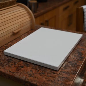 May include: A white rectangular chopping board sits on a granite work surface. A wooden bread bin is to the left. The work surface is reddish-brown. A kitchen scene.