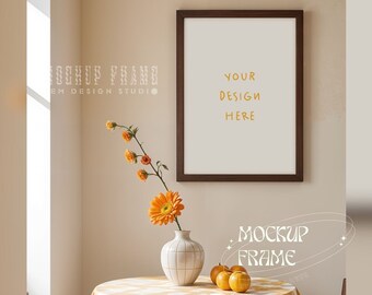 May include: A framed print with the text "YOUR DESIGN HERE" hangs on a beige wall. A round table with a yellow and white checkered tablecloth holds a white vase with orange flowers and small pumpkins. The words "MOCKUP FRAME" are also visible.