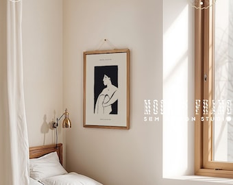 May include: A framed art print featuring a black and white illustration of a seated figure. The artwork is displayed in a gold frame and hangs above a bed with white linens. A brass wall lamp and a window with natural light are also visible.