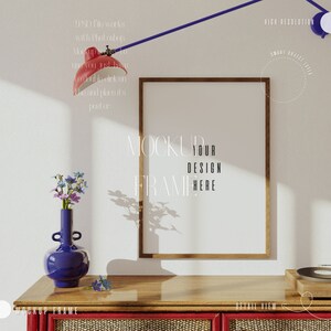 May include: A wooden frame mockup with a blank white space inside the frame. The frame is on a wooden table with a blue vase with flowers and a record player on the table. The text "MOCKUP YOUR DESIGN FRAME HERE" is written on the wall behind the frame.