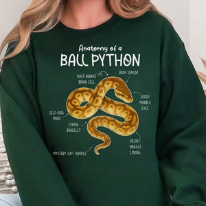 Op de afbeelding: Donkergroene sweatshirt met een gedetailleerde illustratie van een koningspython met gelabelde anatomische kenmerken. De tekst "Anatomy of a BALL PYTHON" staat boven de illustratie van de slang. De sweatshirt is een casual stijl.