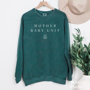 May include: Teal-colored sweatshirt with the text "MOTHER BABY UNIT" in white, and a small teddy bear graphic. The long-sleeved crewneck top is hanging on a wooden hanger. A neutral-toned hat and greenery are in the background.