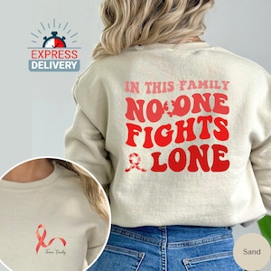 Op de afbeelding: Zandkleurige trui met de tekst "In this family no one fights alone" in rode en roze letters. Een roze lint bevindt zich in de linkerbenedenhoek van de trui. De trui wordt gedragen door iemand die een blauwe jeans draagt.