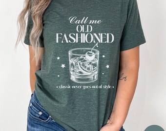 Puede incluir: Una camiseta verde con el texto "Call me OLD FASHIONED" y un dibujo de un cóctel old fashioned. El texto debajo del dibujo dice "classic never goes out of style."