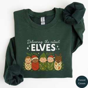 Puede incluir: Sudadera verde bosque con el texto "Delivering the cutest ELVES" e ilustraciones de cinco bebés elfos. Los elfos llevan gorros y están envueltos en mantas estampadas, ideal para la temporada navideña.
