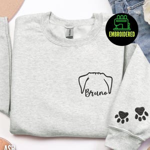 May include: Light gray sweatshirt with a black embroidered dog face and the name "Bruno" below it. Two black paw prints are embroidered on the sleeve. The sweatshirt has a crew neck and is made of a soft material.