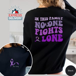 Puede incluir: Sudadera negra con una cinta morada y el texto "En esta familia nadie lucha solo".