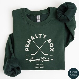 May include: Forest green sweatshirt with a white graphic of crossed hockey sticks inside a circle with the text "Penalty Box Social Club" and "Est 2025".