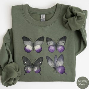 May include: Military green sweatshirt featuring four butterfly illustrations with purple, white, and gray wings. The butterflies are arranged in a square pattern on the front of the long-sleeved crew neck top. The sweatshirt is made of a soft material.