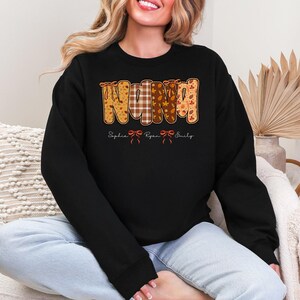 Op de afbeelding: Zwart sweatshirt met het woord "NANA" in een herfstontwerp. De letters zijn versierd met patronen van bladeren, ruiten en bloemen. Daaronder staan de namen "Sophia", "Ryan" en "Emily" gedrukt.