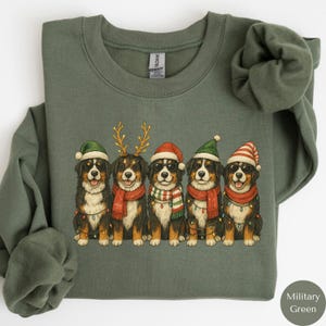 May include: Military green sweatshirt featuring a festive design of five Bernese Mountain Dogs. Each dog is wearing holiday accessories like Santa hats, antlers, and scarves. The dogs are also wearing sunglasses and are adorned with string lights.