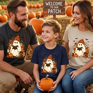 May include: Three t-shirts featuring a graphic of a ghost wearing an orange beanie, reading a book, surrounded by sunflowers, pumpkins, and bats. The adult shirts are charcoal and beige, while the child's shirt is navy blue, all with identical prints.