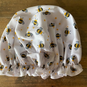 May include: White shower cap with a bee pattern. The bees are yellow and black with small white wings. The cap has a ruffled edge.