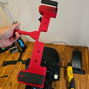 May include: A red and black 3D-printed device, possibly a component for a flight simulator, held in a hand. The device has a T-shape with a black bracket and is positioned over a control panel. A yellow and black DeWalt screwdriver is also visible.