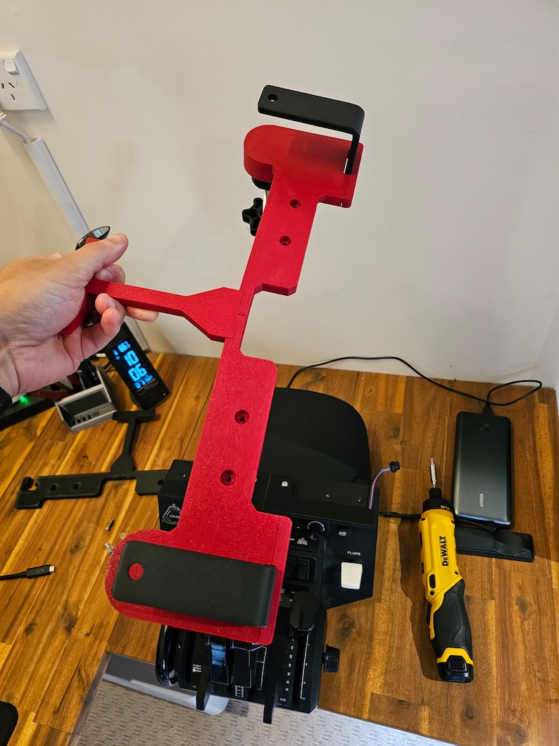 May include: A red and black 3D-printed device, possibly a component for a flight simulator, held in a hand. The device has a T-shape with a black bracket and is positioned over a control panel. A yellow and black DeWalt screwdriver is also visible.
