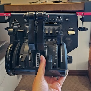 May include: Black flight simulator throttle quadrant with multiple levers, dials, and switches. The unit is mounted to a wooden surface with red and black clamps. The word "GEAR" is visible on the panel.