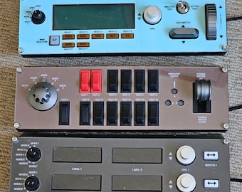 May include: Three rows of electronic control panels with various buttons, knobs, and switches. The top panel is light blue with a digital display and the text "TOGGLE SWITCH", "INACTIVE TEXT", "ACTIVE TEXT", and "JOG SWITCH". The middle panel is brown with the text "TEXT 1" through "TEXT 13", "INDICATOR LIGHTS", and "TOGGLE SWITCH DOWN". The bottom panel is brown with the text "MODE 1" through "MODE 7", "LABEL 1", "LABEL 2", "DIAL 1", "SWITCH 1", "DIAL 2", and "SWITCH 2".