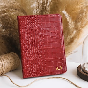 Op de afbeelding: Een rode, getextureerde leren journalhoes met een krokodillenpatroon. De hoes heeft gouden initialen "A.Y" in de rechter benedenhoek. Het tijdschrift wordt gepresenteerd met decoratieve gedroogde planten en een decoratieve glazen koepel.