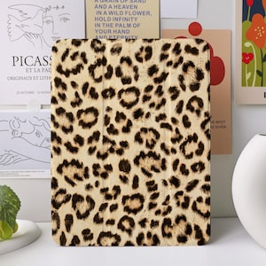 Puede incluir: Una funda para tableta con estampado de leopardo. La funda tiene un fondo beige con manchas marrones oscuras. La funda rectangular está hecha de un material duradero. La funda se muestra frente a un jarrón blanco y obras de arte.
