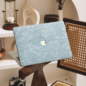 May include: A light blue laptop with a floral design is propped on a wooden chair. The laptop has a silver Apple logo. The background includes a vintage camera, hourglass, and decorative vases, suggesting a home decor theme.