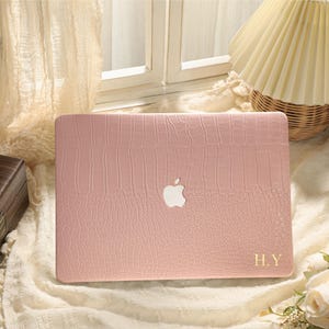 May include: A pink laptop case with a crocodile skin texture. The case features the Apple logo and gold initials "H.Y." The case is on a white surface with a window and a lamp in the background.