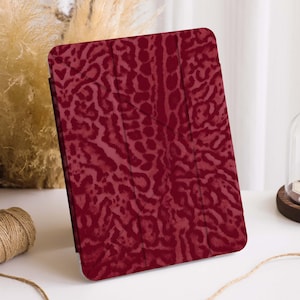 Puede incluir: Una funda de tableta burdeos con un patrón repetitivo que se asemeja a un estampado animal. La funda está abierta, revelando el interior. El fondo incluye elementos decorativos.