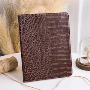May include: A brown, faux-leather tablet case with a crocodile skin texture. The case is rectangular and closed, with a stitched border. The background includes dried plants, twine, and a decorative glass dome.