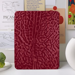 Könnte beinhalten: Eine weinrote Tablethülle mit einem strukturierten Animal-Print-Design. Die rechteckige Hülle hat abgerundete Ecken. Der Hintergrund umfasst Kunstdrucke und eine weiße Vase.