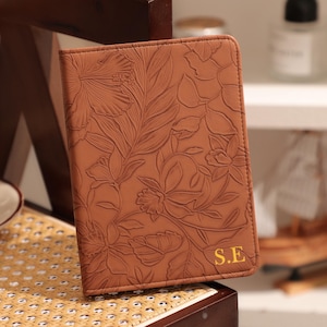 May include: A brown leather journal with an embossed floral design. The cover features a detailed pattern of flowers and leaves. The initials "S.E" are embossed in gold on the lower right corner. The journal is closed and appears to be new.