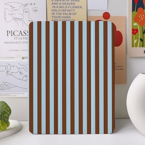 May include: A rectangular object with vertical brown and light blue stripes. It is positioned in front of a white vase and several art prints. The art prints feature text and illustrations. One print says "BLOSSOM".