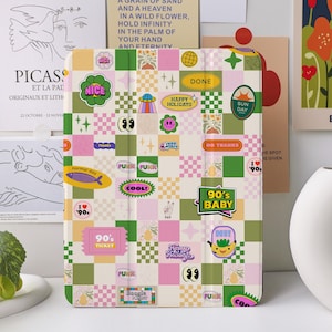 Puede incluir: Una funda blanca para iPad con un patrón a cuadros en rosa, verde y blanco. La funda está cubierta de pegatinas coloridas con diseños de temática de los 90, incluyendo "90's Baby", "Cool", "Felices Fiestas" y "Día de Sol".