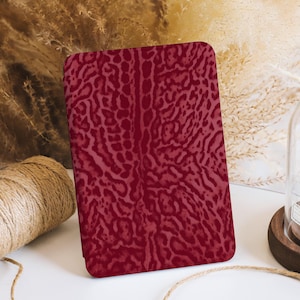 Puede incluir: Un cuaderno de color burdeos con un diseño texturizado de estampado animal. El cuaderno rectangular está de pie sobre una superficie blanca. El fondo incluye plantas secas y una bobina de hilo.