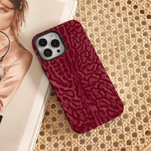 Könnte beinhalten: Eine weinrote Handyhülle mit einem strukturierten Animal-Print-Design. Die Hülle hat einen schwarzen Rand um die Kamera und liegt auf einer gewebten Oberfläche.