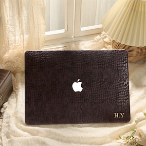 Könnte beinhalten: Eine dunkelbraune Laptop-Hülle mit einer strukturierten, reptilienartigen Musterung. Das Apple-Logo ist sichtbar, zusammen mit den goldenen Initialen "H.Y.". Die Hülle liegt auf einer cremefarbenen Oberfläche, mit einem Fenster und einer Lampe im Hintergrund.