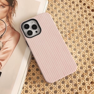 Puede incluir: Una funda de teléfono a rayas rosas y blancas. La funda está hecha de un material duro y tiene un contorno negro para la lente de la cámara. La funda se muestra sobre una superficie tejida.