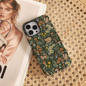 May include: A green phone case with a floral pattern featuring cats, mushrooms, and flowers. The case is designed for an iPhone with a camera lens cutout.