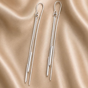 May include: A pair of long silver drop earrings featuring a hook closure. Each earring consists of two thin, textured metallic chains hanging vertically, accented by two smooth, cylindrical silver bars of varying lengths at the bottom.