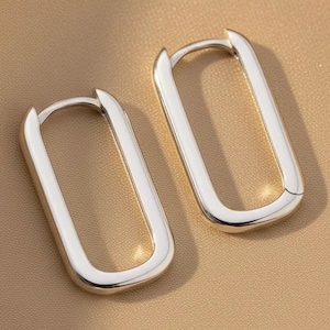 Peut inclure: Une paire de boucles d'oreilles créoles argentées présentant une forme rectangulaire allongée aux coins arrondis. Les boucles d'oreilles ont une finition métallique lisse et polie ainsi qu'un mécanisme de fermeture à charnière.