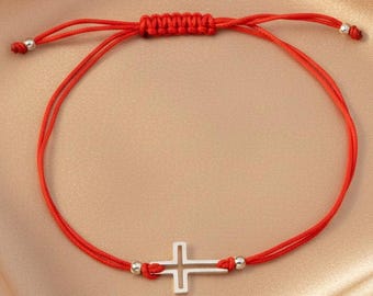 May include: A red string bracelet featuring a silver-toned metal cross charm at the center. The bracelet includes an adjustable sliding knot closure and two small silver-toned beads at the ends of the cord, creating a simple and clean design.