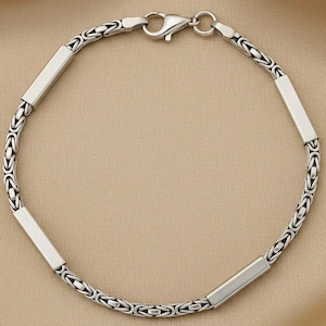 May include: A silver chain bracelet featuring a repeating Byzantine link pattern interspersed with four smooth, polished rectangular silver bars. The bracelet is secured with a silver lobster claw clasp, creating a circular shape.