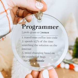 May include: A clear, round ornament with the word "Programmer" and its definition printed in black. The definition includes humorous statements about coding. A gold-colored string is attached for hanging.