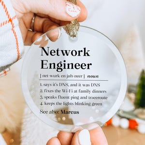 May include: A round, clear glass ornament featuring black printed text that defines a Network Engineer with four humorous points, including fixing Wi-Fi and speaking fluent ping, finished with a gold ribbon loop for hanging on a tree.