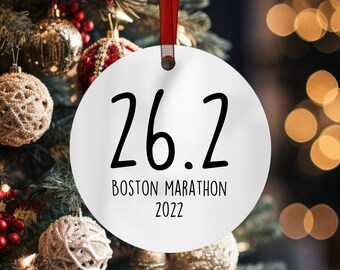 May include: A white, round Christmas ornament with the text "26.2 Boston Marathon 2022" in black. The ornament hangs from a red ribbon and is surrounded by a Christmas tree with ornaments and lights.