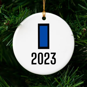 May include: White ceramic Christmas ornament with a blue rectangle and the year 2023 in black. The ornament hangs from a gold string against a backdrop of green Christmas tree branches.