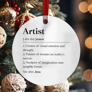 May include: A white, round Christmas ornament with the definition of "Artist" printed on it. The definition includes the pronunciation, the word "noun", and three descriptive statements. The ornament hangs from a red ribbon.